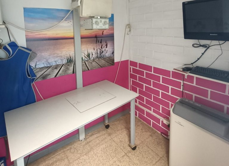 Sala con una mesa de exploración, un monitor, un aparato de rayos x, y un traje protector de irradiación.